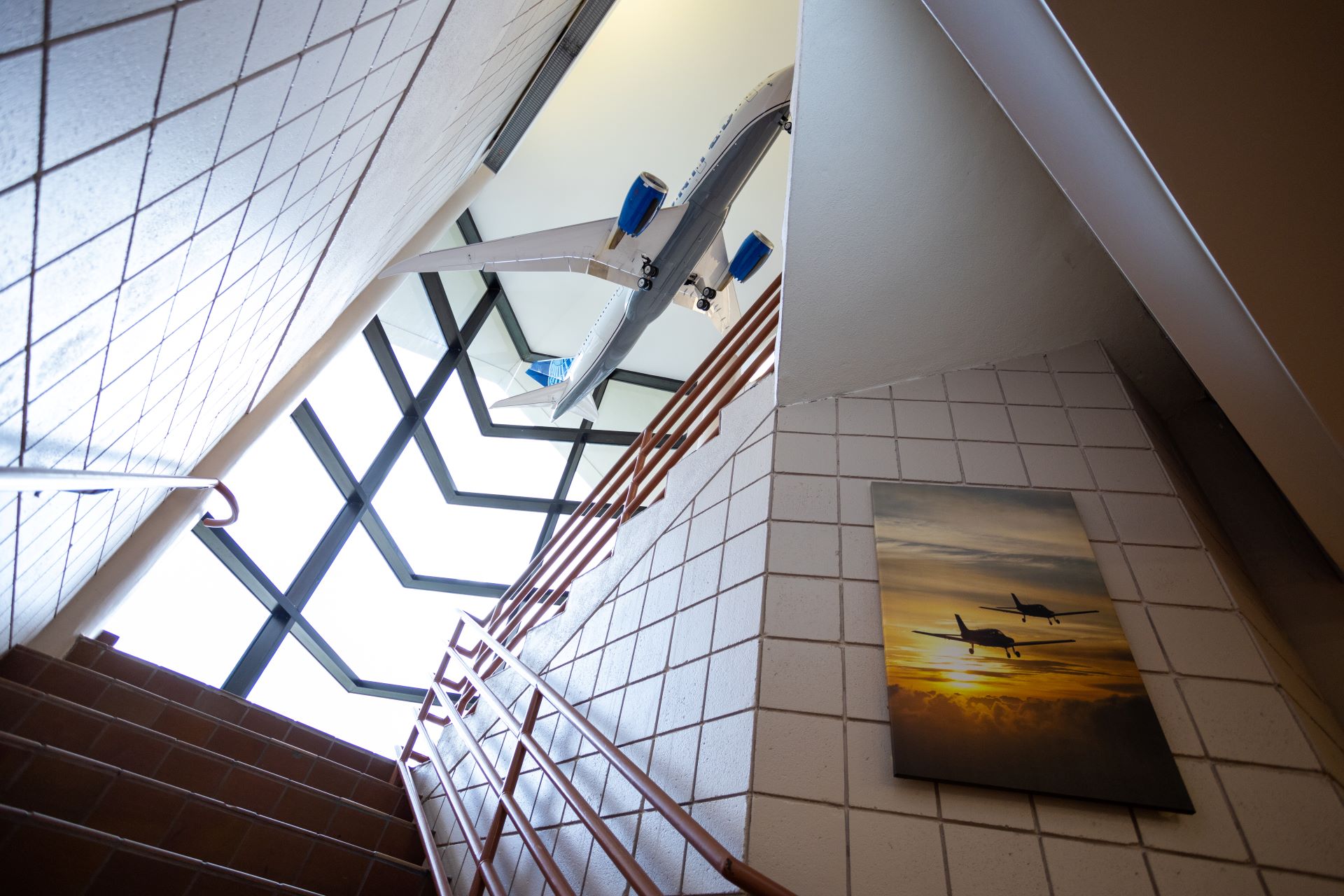 Upward view of a stairwell with a model airplane suspended above, next to a geometric window and a wall-mounted picture of an airplane flying at sunset